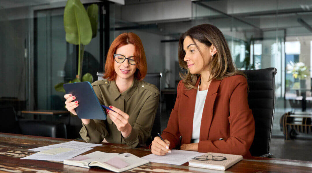 Two busy business women talking at desk in office.