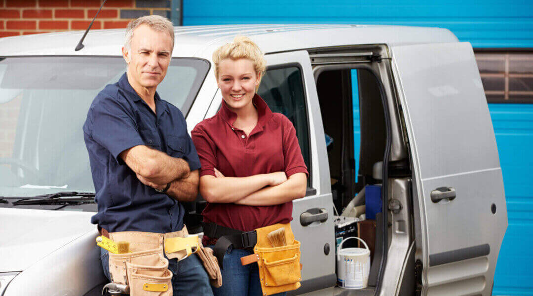 Mature man wearing tool belt standing next to a younger woman, also wearing a tool belt, in front of a work van smiling at camera