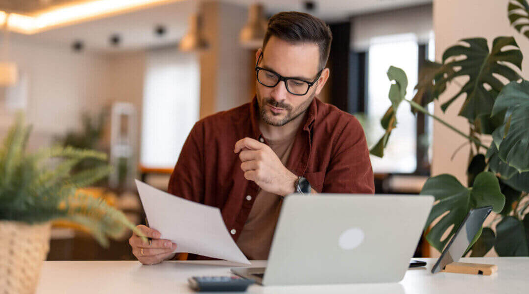 man using laptop sitting at the table in a home office, focused guy in casual clothing looking at the paper, communicating online, writing emails, working.