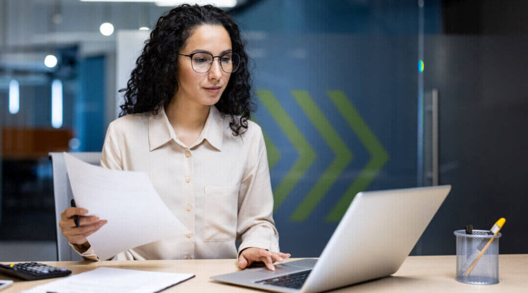 Pensive business woman works inside office, holding a piece of paper and looking at a laptop screen, thinking about contracts and reports with charts and graphs