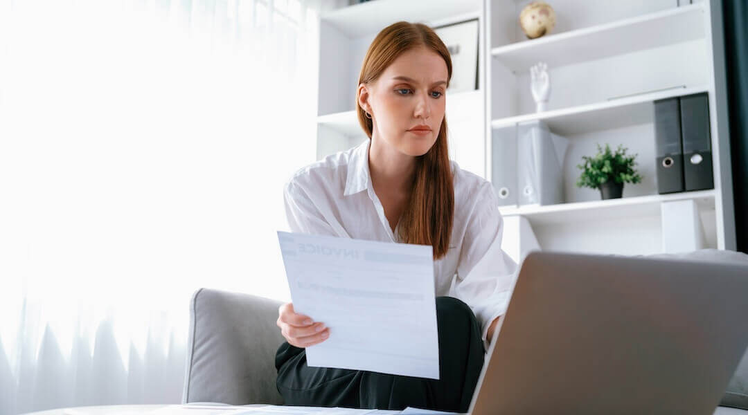 Entrepreneur compares financial statement on her laptop to a printed statement she holds in her hand while sitting in an office